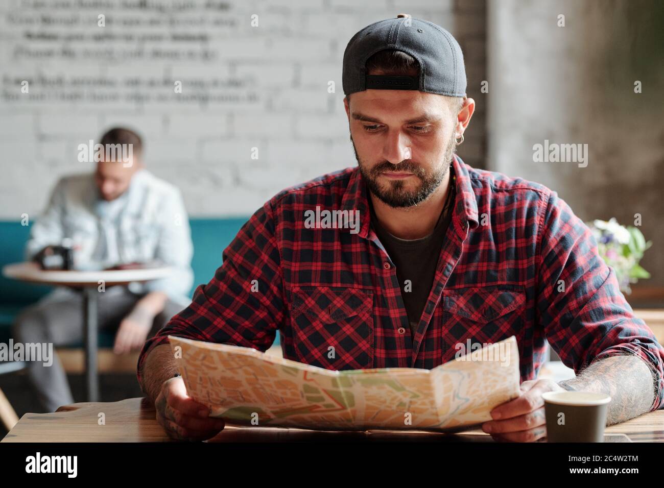 Konzentrierter junger bärtiger Mann in einer Ballkappe, der am Tisch sitzt und im Café Papierkarte verwendet Stockfoto