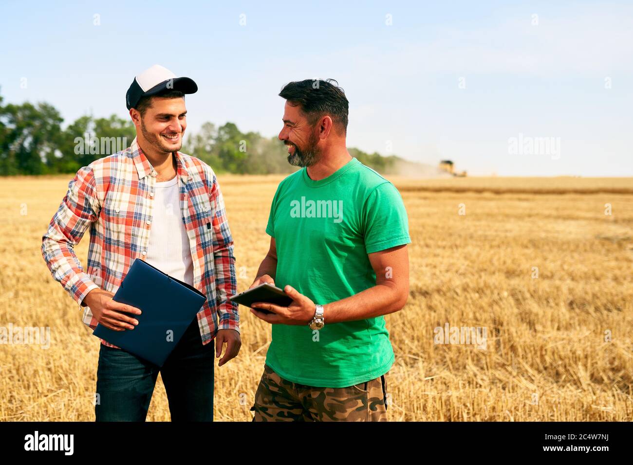 Zwei Bauern stehen im Weizenstoppelfeld, diskutieren Ernte, Ernte. Senior Agronom mit Touch Tablet pc lehrt junge Mitarbeiter. Innovative Technik Stockfoto