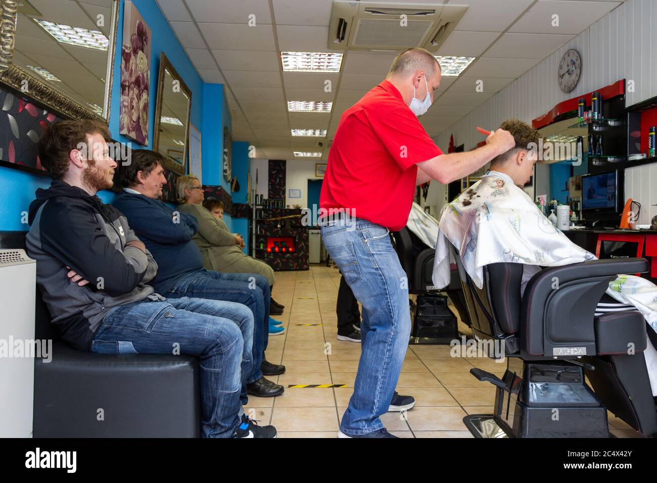 Barber trägt eine Gesichtsmaske, die einem mann in einem Friseurladen Haare schneidet, während Männer für einen Haarschnitt Schlange stehen Stockfoto