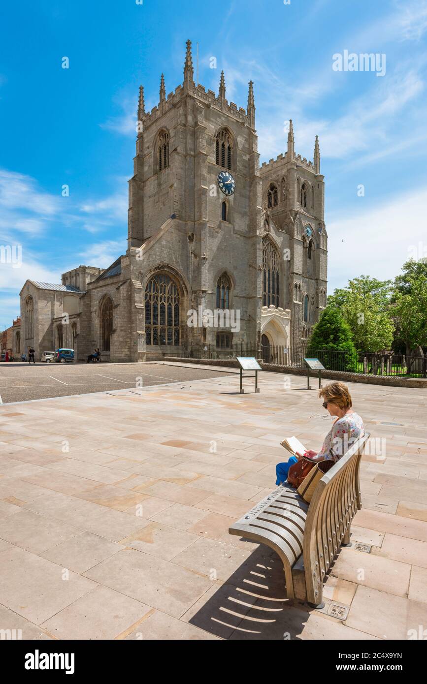 Frau liest Buch Konzept, Blick im Sommer von einer reifen Frau sitzen allein auf einer Bank in der Nähe einer Kathedrale in einer europäischen Stadt, King's Lynn, Großbritannien Stockfoto