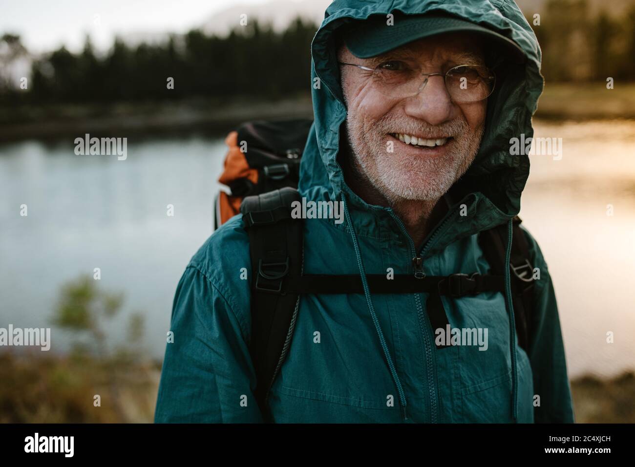 Porträt eines älteren Mannes, der einen Rucksack trägt und die Kamera anschaut und lächelt. Fit alten Mann auf eine Wanderung mit Fluss im Hintergrund. Stockfoto