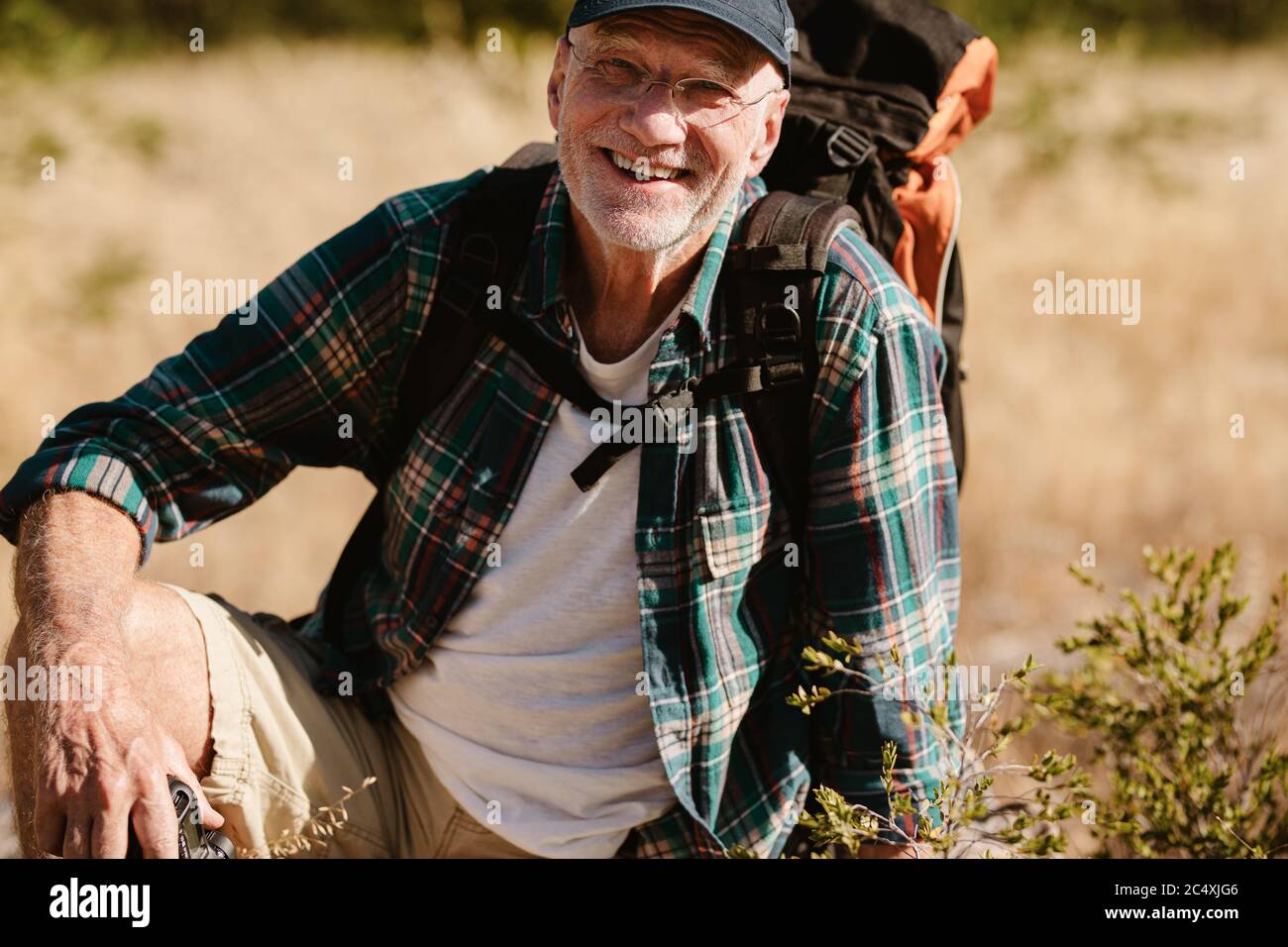 Porträt eines älteren Mannes, der einen Rucksack trägt und die Kamera anschaut und lächelt. Pensionierter männlicher Wanderer, der eine Pause macht, auf dem Boden sitzend. Stockfoto