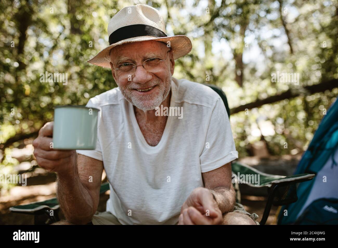 Lächelnder älterer Mann mit erfrischendem Kaffee auf dem Campingplatz. Männlicher Reisender hält eine Tasse Kaffee und schaut auf die Kamera beim Campingausflug. Stockfoto