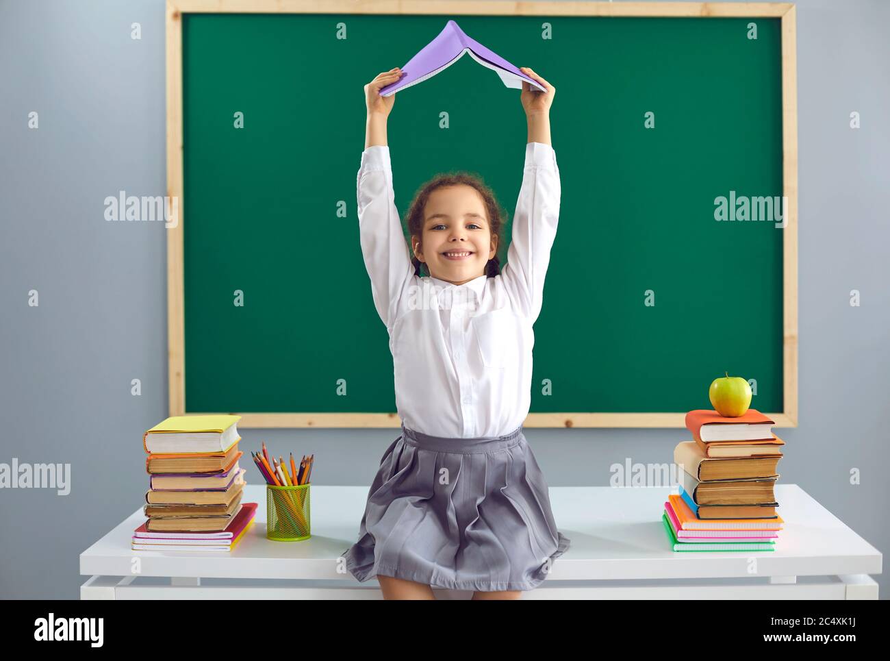 Zurück zur Schule. Fröhliches kleines Mädchen, das auf dem Schreibtisch sitzt und das Buch über ihrem Kopf im Klassenzimmer hält. Nettes Kind Spaß in der Schule Stockfoto