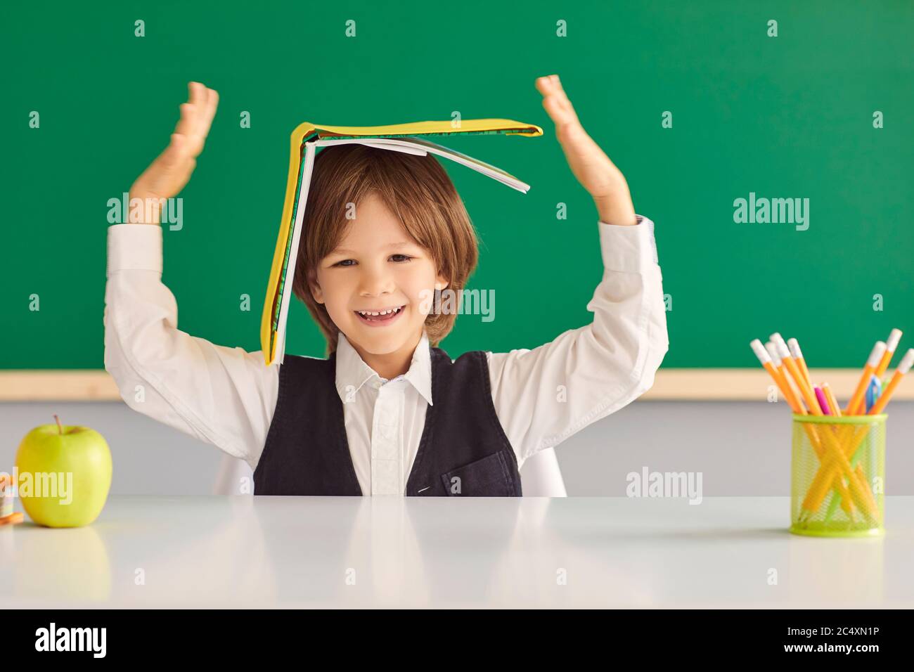 Zurück zur Schule. Fröhlicher kleiner Junge versteckt unter Buch, spielen dummes Spiel am Schreibtisch im Klassenzimmer. Stockfoto