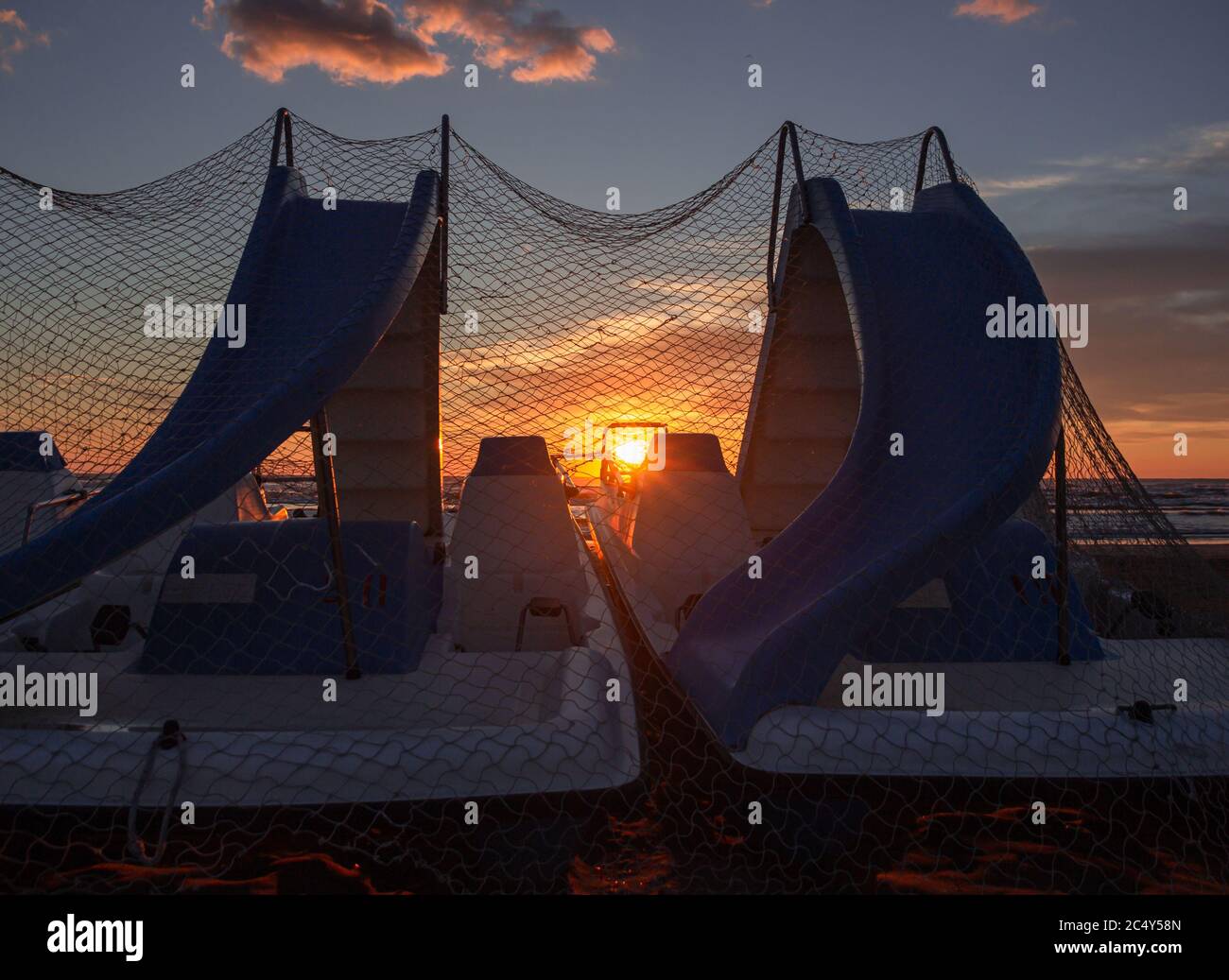 Sonnenstrahlen, die sich auf Tretbooten spiegeln, die mit Fischernetzen bedeckt sind, am Morgen während des wunderschönen Sonnenaufgangs am Strand von Rivazzurra (Rimini/Italien) Stockfoto