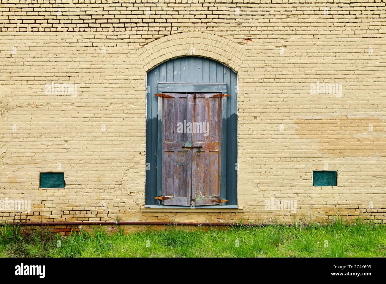 Ein lackiertes gelbes Ziegelgebäude mit vertaufter grüner Eingangstür und Grasfront mit rostigen Rohren Stockfoto