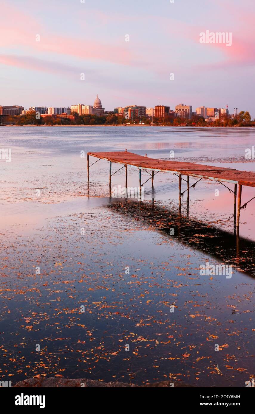 Skyline von Madison in der Innenstadt mit Kuppel des Wisconsin State Capitol Gebäudes, die von der anderen Seite der Monona Bucht bei Sonnenuntergang gesehen wird. Hölzerne Pier und gefallene Blätter o Stockfoto