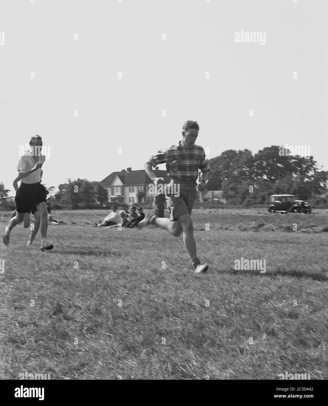 50er Jahre, historische, Erwachsene Männer in kurzen Hosen und langärmeligen Hemden, die an einem Laufrennen draußen auf einem Gras in einem Dorf grün, möglicherweise Scout-Meister und Senior Scouts im Scouting Sommerlager. Ein Auto der Zeit kann in der Ferne geparkt gesehen werden. Stockfoto
