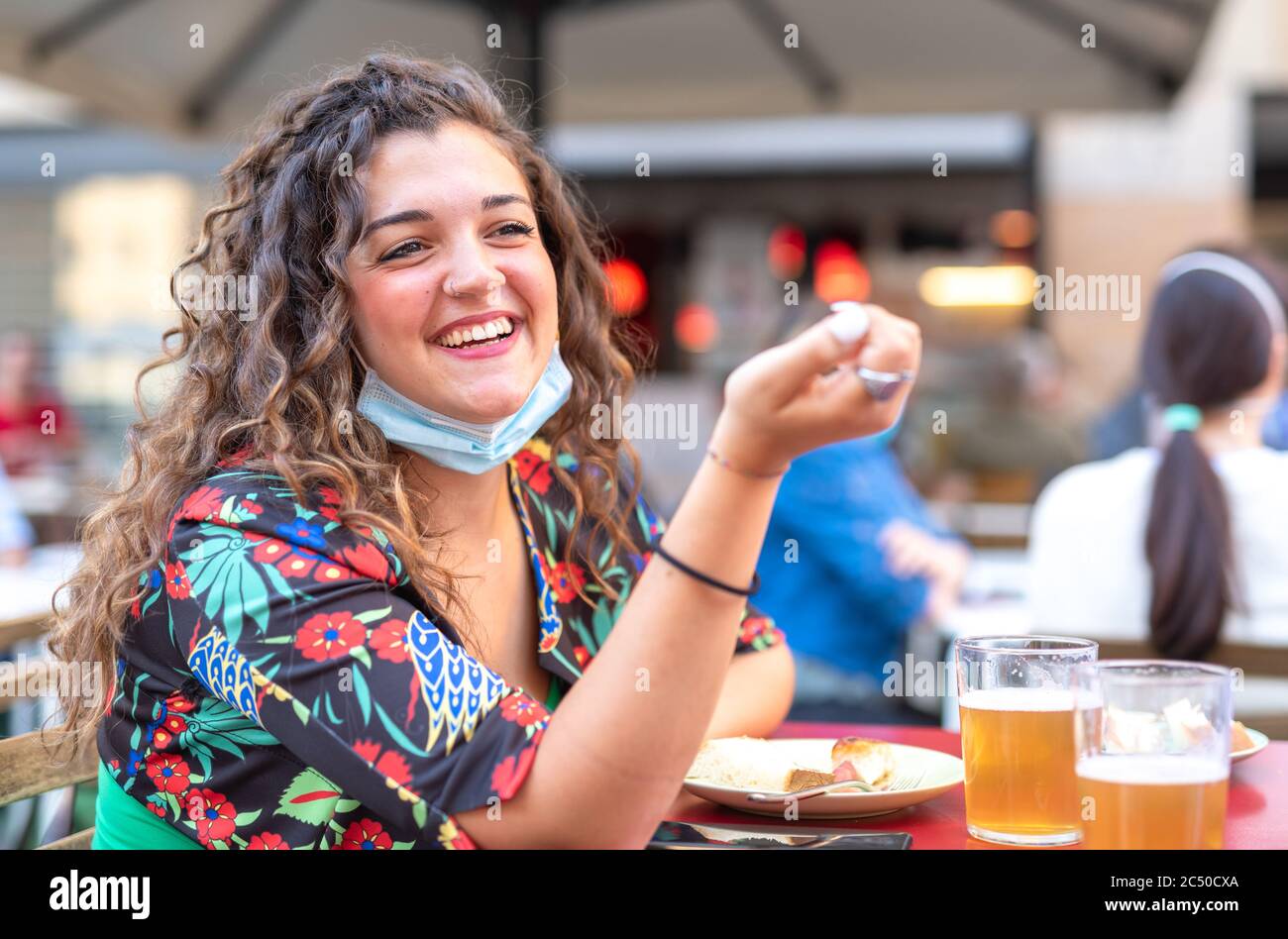 Junge Mädchen mit lockigen Haaren, die lacht, während sie Bier in einer Bar trinken, trägt eine Schutzmaske unter ihrem Kinn, Konzept der Wiederaufnahme der sozialen aktiv Stockfoto