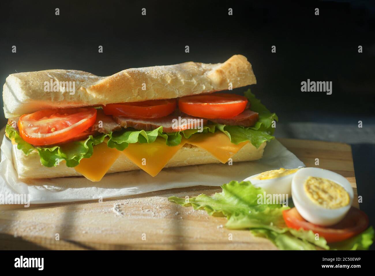 Ein appetitliches Sandwich mit Schinken, Käse, Tomaten und Salat liegt auf einem Brett mit Ei und Zitrone auf schwarzem Hintergrund Stockfoto