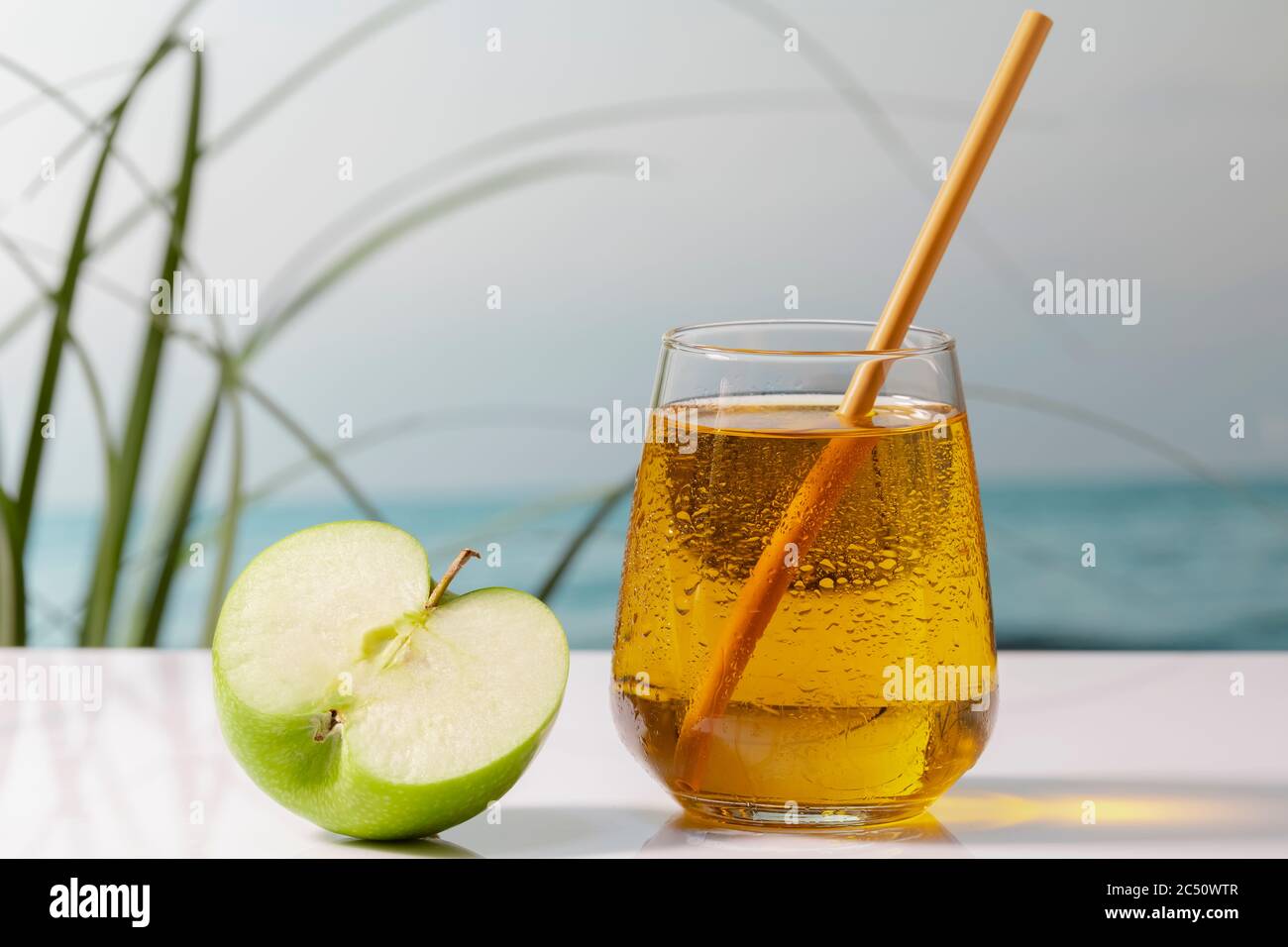 Nahaufnahme eines verlockenden kalten Glases Apfelsaft mit einem Stroh neben einem halben Apfel auf einem Tisch auf einem unscharf Hintergrund. Urlaub und veganes Essen con Stockfoto