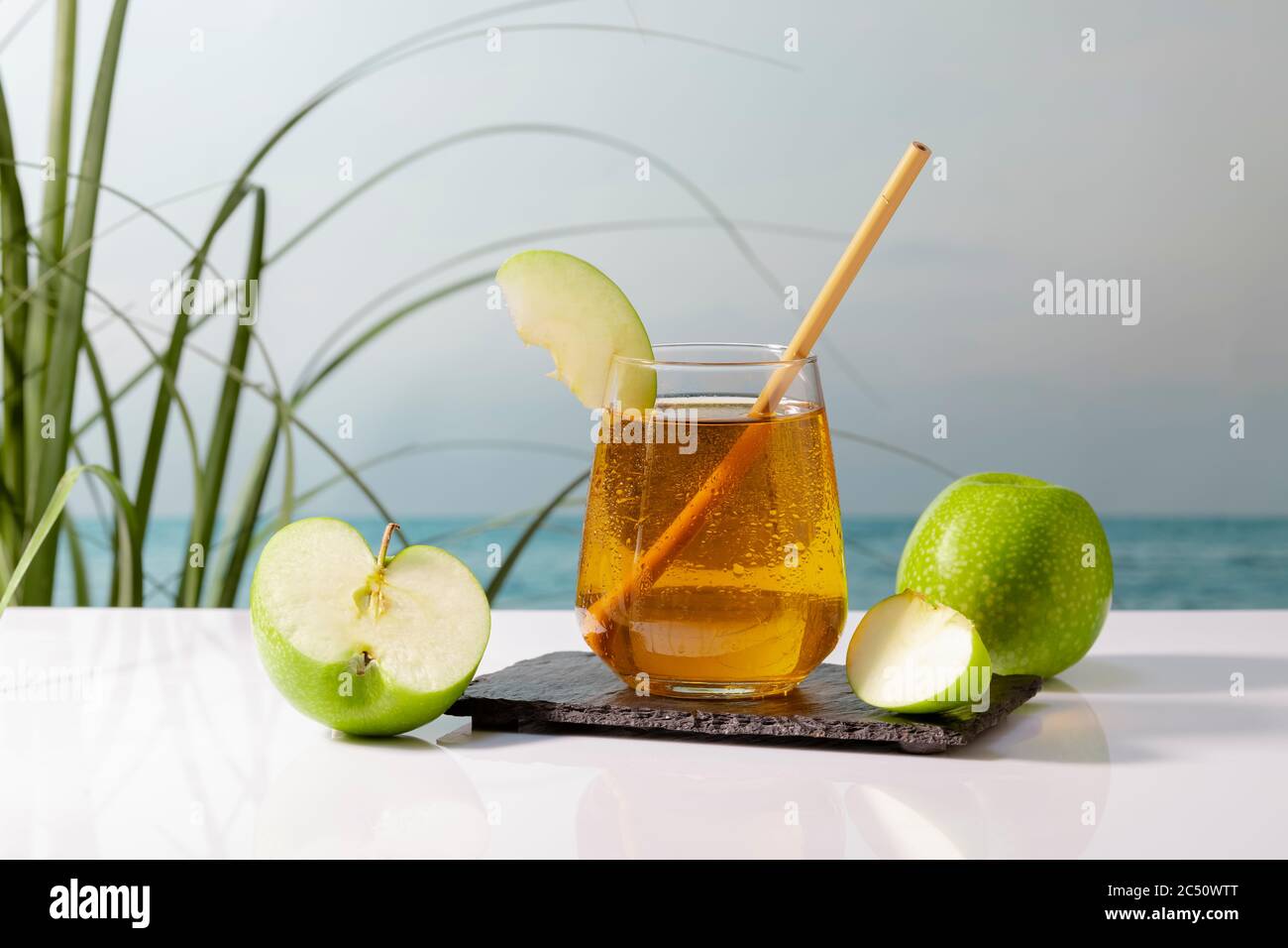 Ein köstlich aussehendes Glas Apfelsaft mit einem Stroh und einer Apfelscheibe auf einem dunklen Teller, umgeben von Äpfeln auf einem unscharf dargestellte Hintergrund. Vegan und h Stockfoto