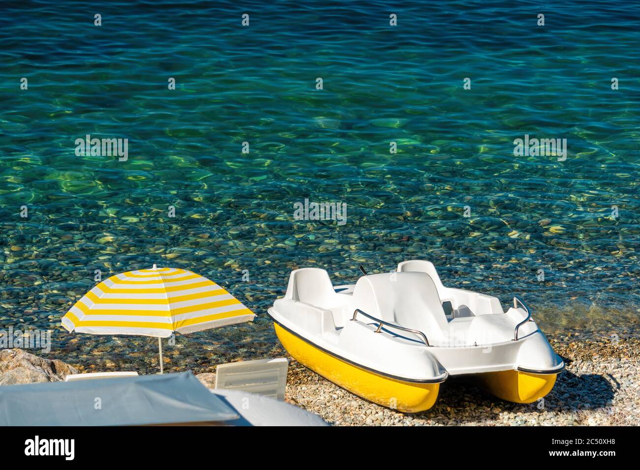 Weißer Katamaran am Kieselstrand mit gelbem Sonnenschirm, azurblaue kristallklare Küste. Speicherplatz kopieren. Das Konzept einer Reise, Entspannung, aktiv und Stockfoto