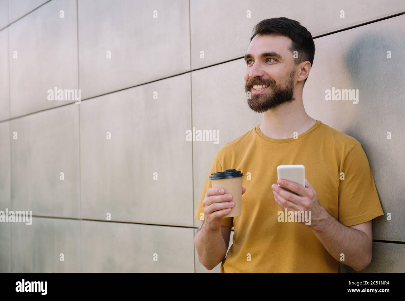 Junge bärtige halten Smartphone und Tasse Kaffee, Online-Shopping. Lächelnder Hipster mit Handy, wartet draußen auf Taxi Stockfoto