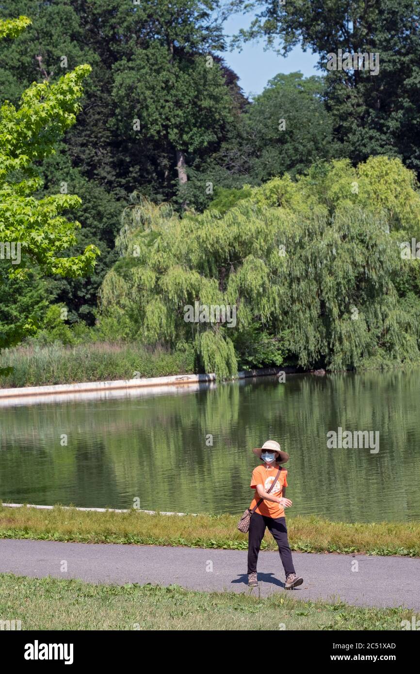 Eine Frau in einem Strohhut und mit einer Maske geht schnell in der Nähe des Sees in Kissena Park, Flushing, Queens, New York City. Stockfoto