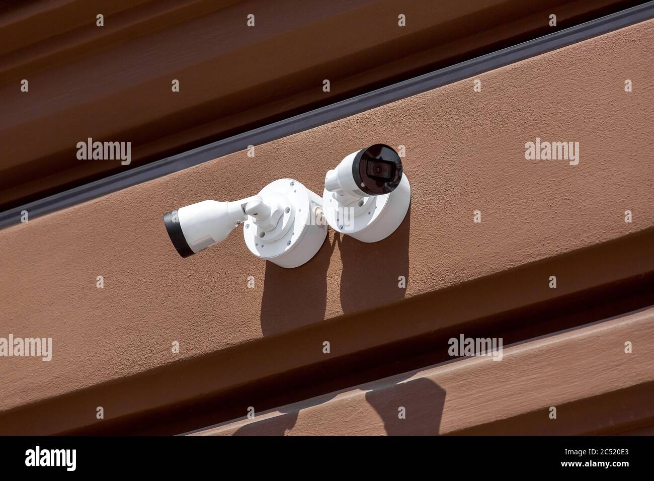 Zwei weiße CCTV-Kameras an der Außenfassade einer braunen Wand, Nahüberwachungssysteme und Fernbedienungskontrolle an sonnigen Tagen. Stockfoto