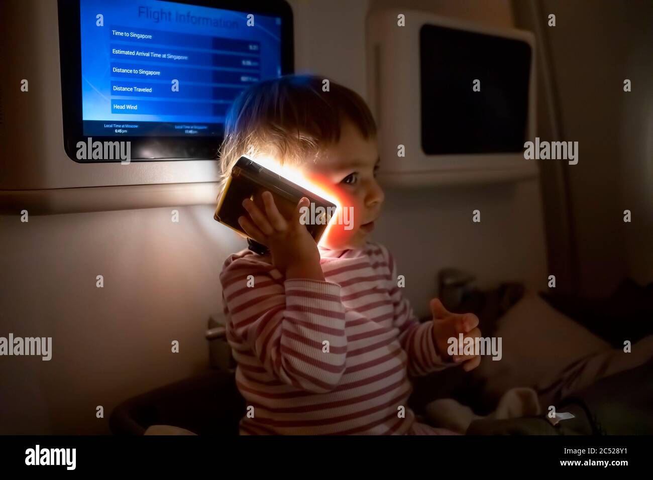 Kleines Kleinkind sitzt in speziellen Babygewanne auf einem Flugzeug in der Dunkelheit und spielt mit einer Taschenlampe Stockfoto