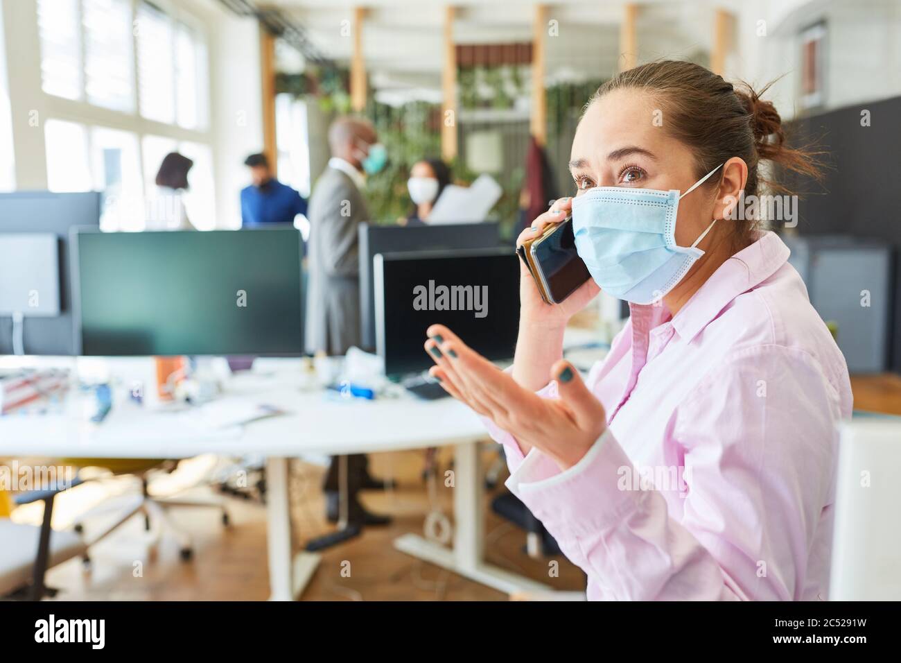 Junge Geschäftsfrau mit Mund-Nase-Schutz im Büro ruft wegen Covid-19 am Handy an Stockfoto