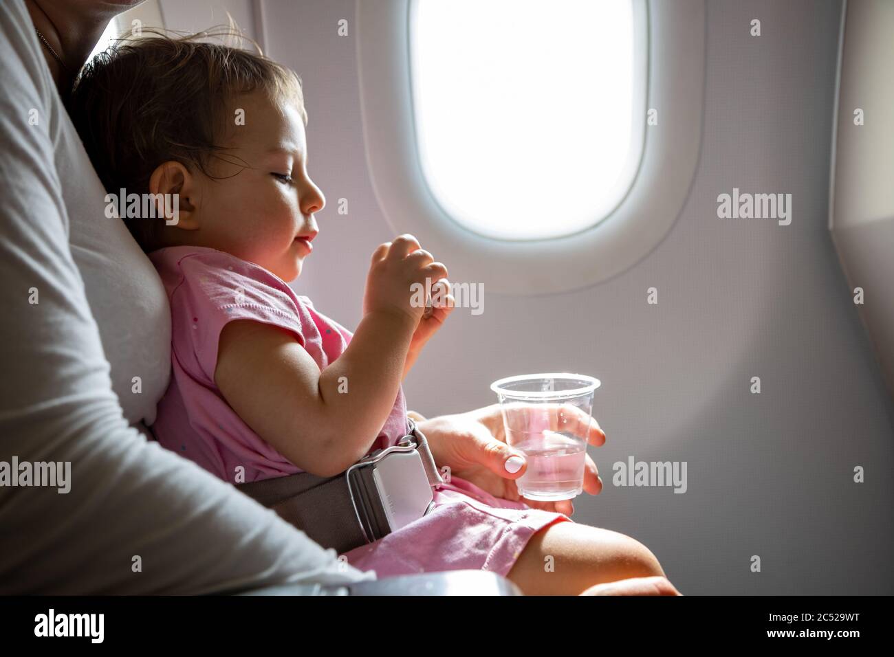 Ein junger Passagierkind, der in einem Flugzeug sitzt und einen Sicherheitsgurt in den Armen seiner Mutter trägt, bereitet sich auf seine erste Reise vor Stockfoto