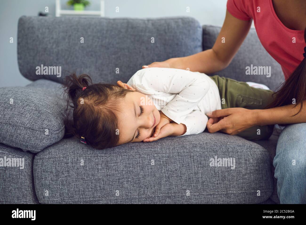 Junge Mutter in der Nähe ihrer niedlichen schlafenden Tochter auf Sofa zu Hause. Eltern beobachten liebliches Kind, das tagsüber ein Nickerchen macht Stockfoto