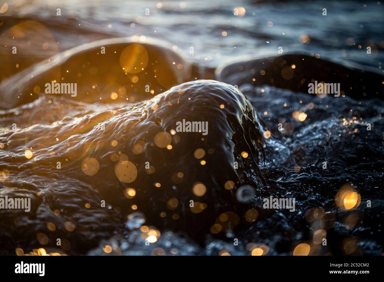 Nahaufnahme von Meereswellen, die auf einer nassen Küste brechen Steine Sommer Sonnenuntergang, Wasserspritzer, Sonnenstrahlen, Reflexionen auf Wassertropfen. Stockfoto