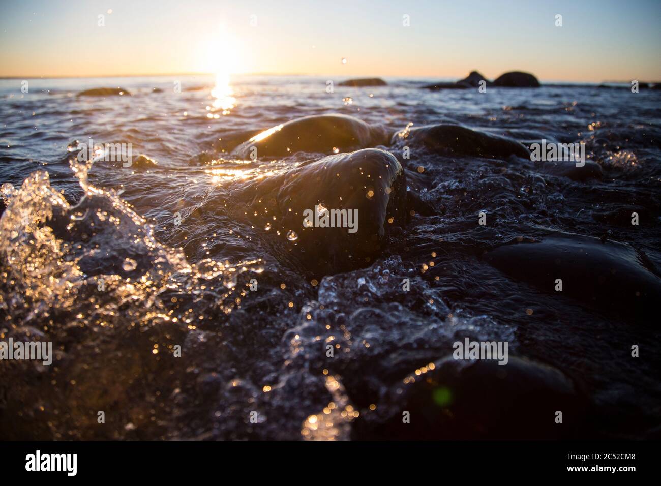 Nahaufnahme von Meereswellen, die auf einer nassen Küste brechen Steine Sommer Sonnenuntergang, Wasserspritzer, Sonnenstrahlen, Reflexionen auf Wassertropfen. Stockfoto