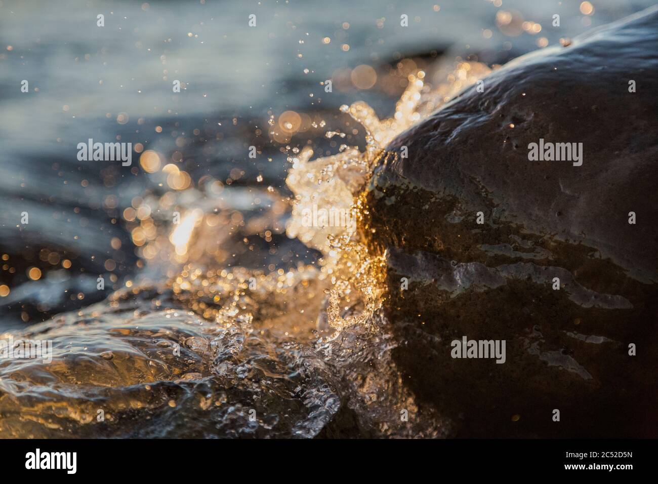 Nahaufnahme von Meereswellen, die auf einer nassen Küste brechen Steine Sommer Sonnenuntergang, Wasserspritzer, Sonnenstrahlen, Reflexionen auf Wassertropfen. Stockfoto