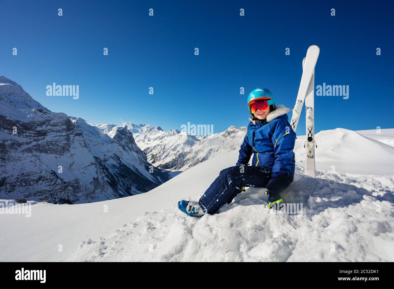 Portrait in den Bergen eines Mädchens in blauem Helm und Skioutfit über dem Himmel sitzen im Schnee auf alpinen Resort Stockfoto