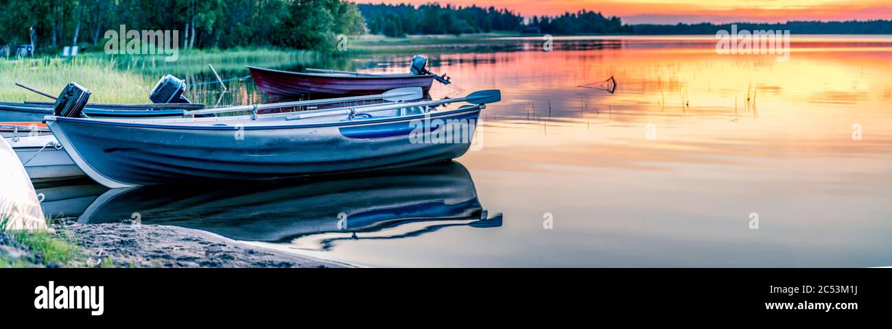 Skandinavisch weiße Nacht in Schweden. Blick auf Motorboote am See. Mitternachtssonne direkt unter der Horizontlinie erleuchten goldene Sonnenstrahlen die höchsten Wolken Stockfoto