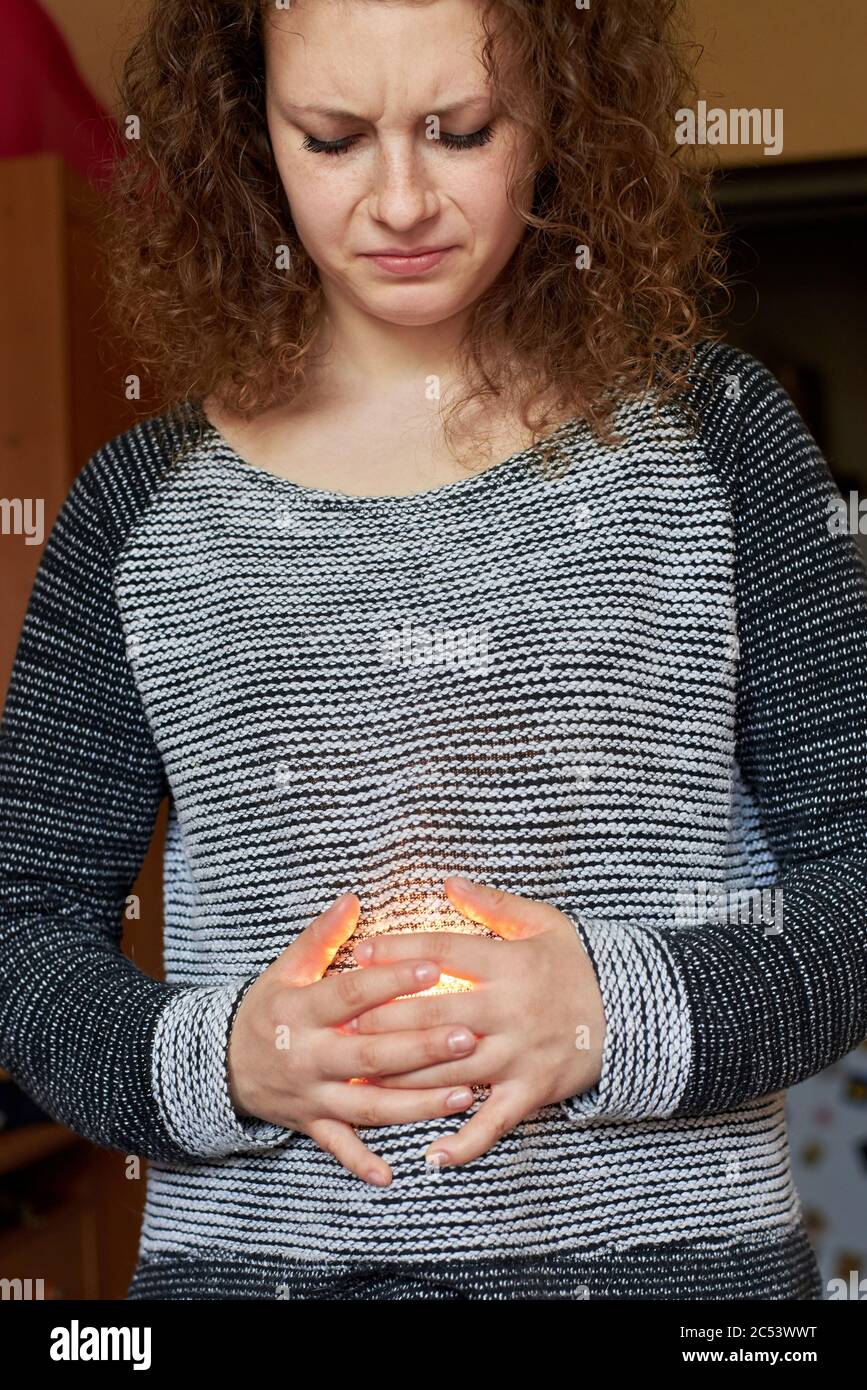 Hübsche Frau mit einem verdrehten Gesicht hält ihre Hände auf ihrem Bauch, durch den das Licht geht. Magenschmerzen Stockfoto