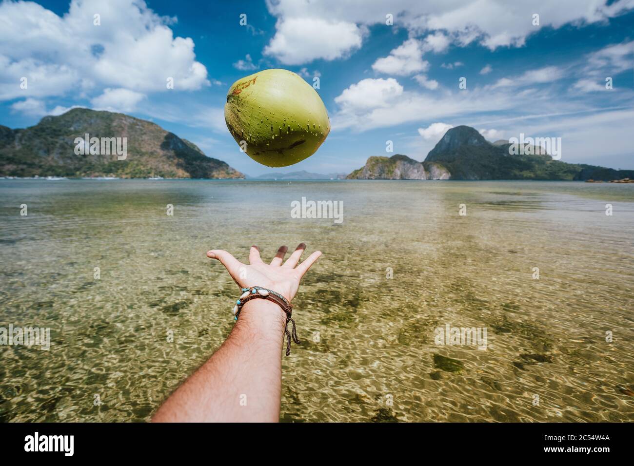 POV jongliert mit Kokosnuss in männlichen Händen gegen exotische Inseln in der Meeresbucht. Stockfoto