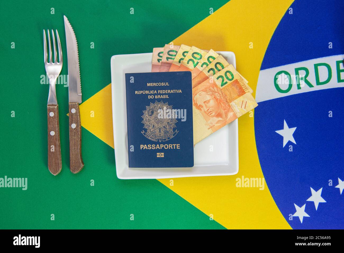 Brasilianischer Reisepass mit echten Banknoten auf einem weißen Teller mit der Flagge Brasiliens im Hintergrund. Besteck neben dem Teller. Stockfoto