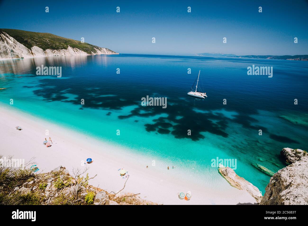 Weiße Katamaran-Yacht in klarem blauem Meerwasser. Touristen am Strand in der Nähe der azurblauen Lagune. Kefalonia, Griechenland. Stockfoto