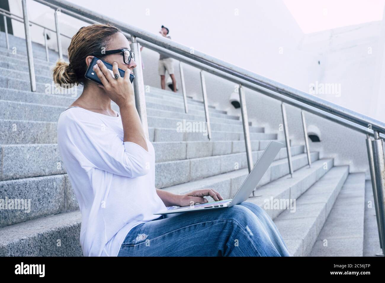 Erwachsene kaukasische Frau, die mit Technologie und modernen Geräten wie Telefon und Laptop im Freien auf einer Treppe im städtischen Hintergrund sitzt - Konzept Stockfoto