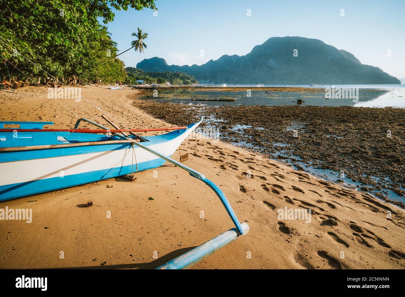 Bangka banca Boot auf sandigen abgelegenen Strand von goldenen Sonnenuntergang Licht beleuchtet. El Nido Dorf im Hintergrund. Philippinen. Stockfoto