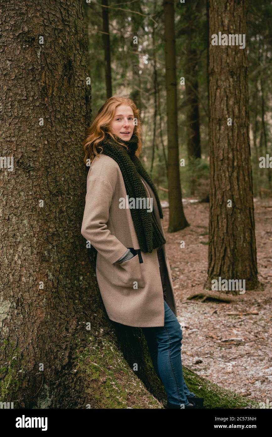 Portrait selbstbewusst Rotschopf Frau in Wollmantel lehnt sich an Baum in Wäldern Stockfoto