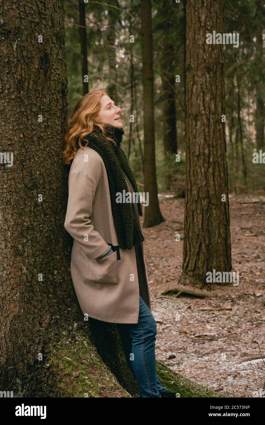 Heitere Rothaarige Frau in Wollmantel und Schal lehnt sich an Baum in Wäldern Stockfoto