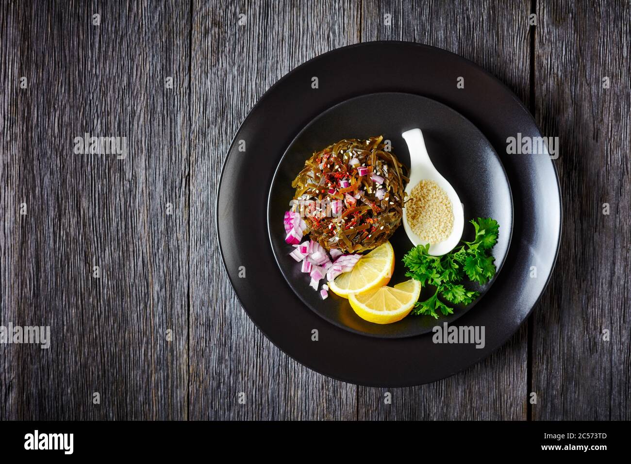 Heißer Algensalat aus eingelegtem Wakame, roten Zwiebeln und Zitronenkeilen, auf einem schwarzen Teller mit frischer Petersilie, bestreut mit Sesamsamen, Chili Flocken auf Stockfoto