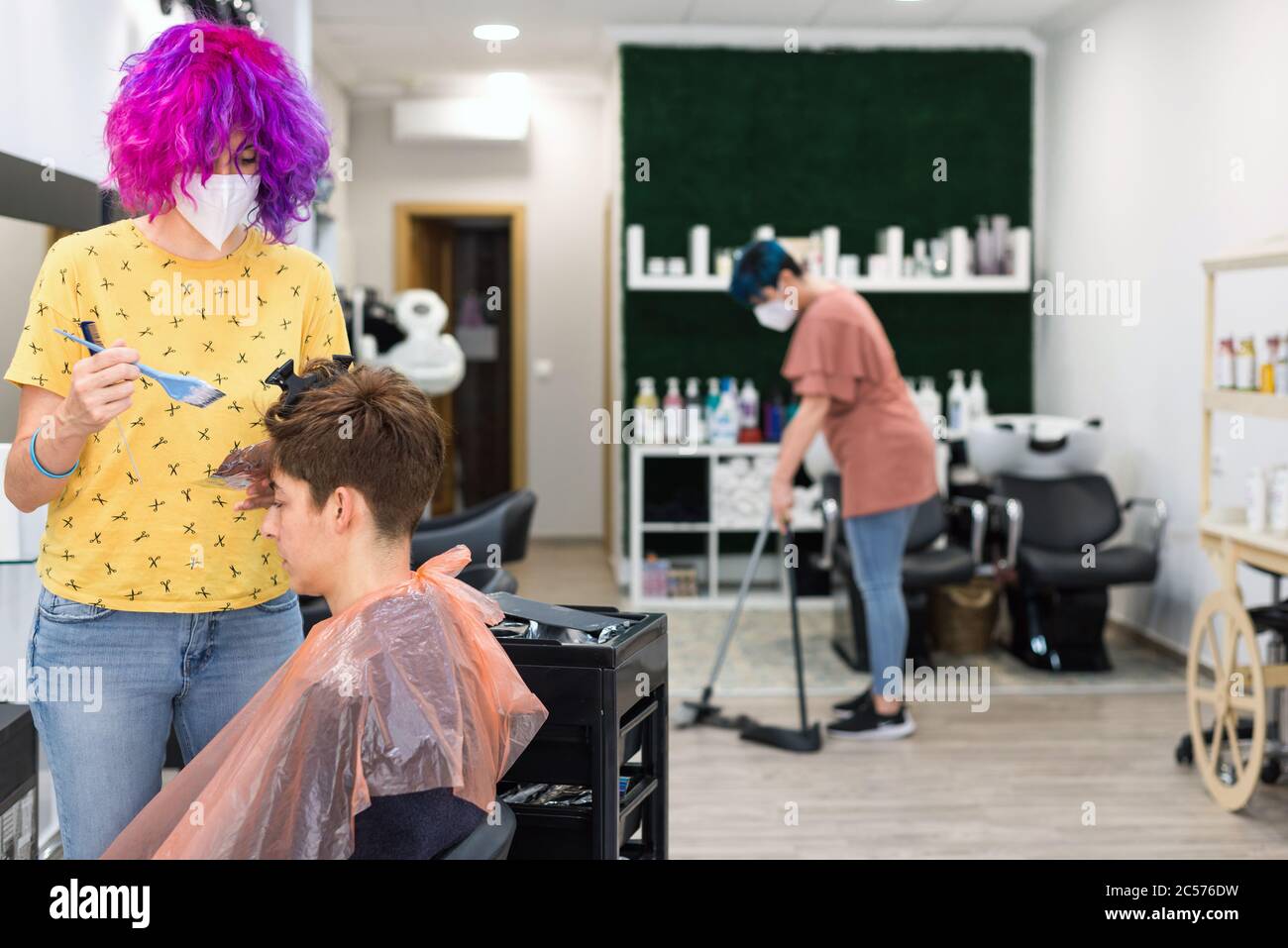 Konzept: Nach dem Coronavirus, covid-19. Professioneller Friseur mit Gesichtsmaske und Handschuhen. Bürsten blonde Highlights. Blauhaarige Frau, die sich fegt Stockfoto