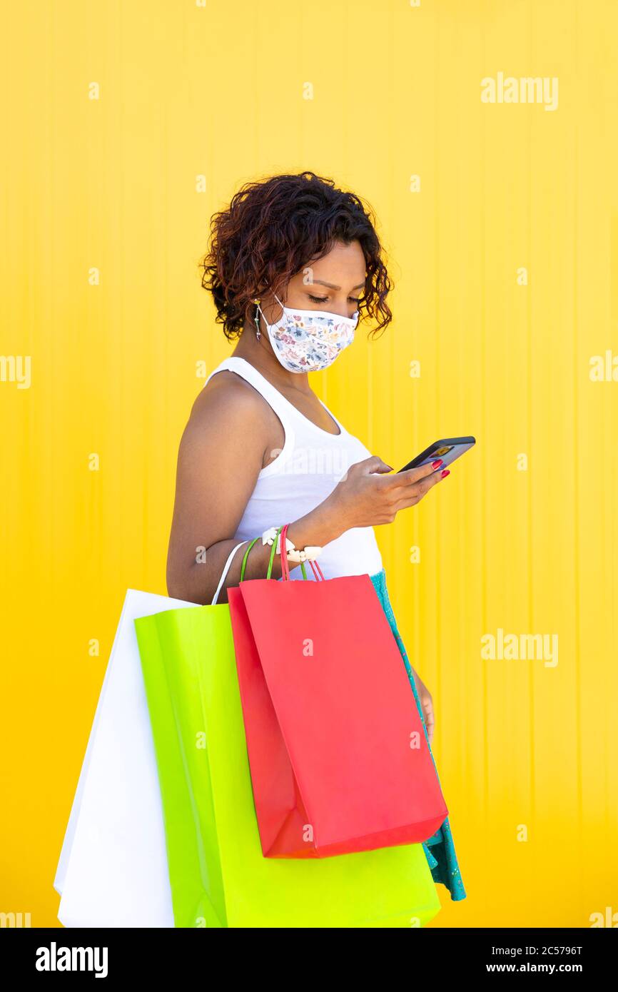 Mädchen mit einer Gesichtsmaske herumlaufen mit ihren Einkaufstaschen und Blick auf ihr Smartphone. Gelber Hintergrund. Leerzeichen für Text. Neues normales Konzept. Stockfoto
