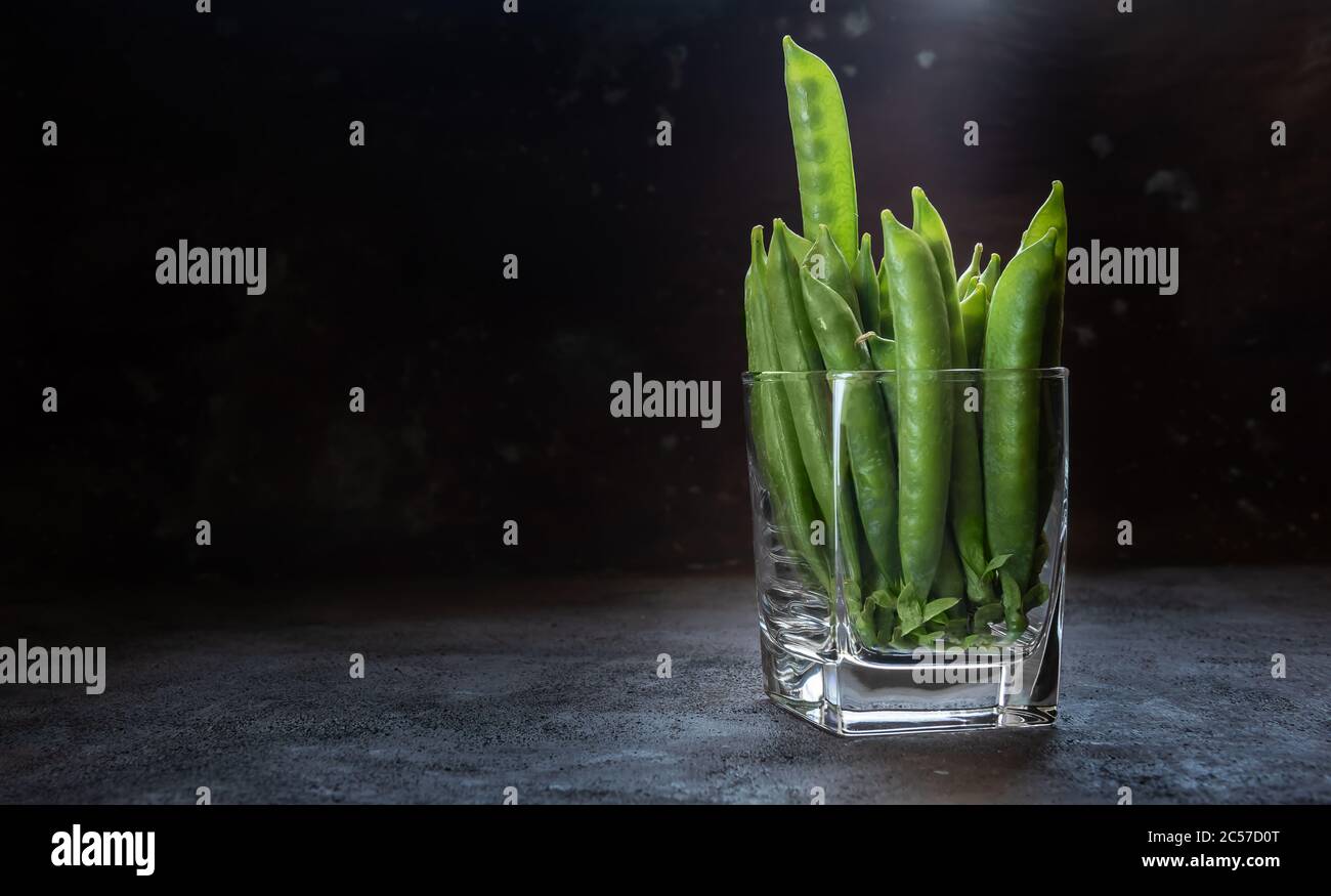 Grüne Erbsen in Glas. Eine Hülse ragt aus dem Stapel und die Samen sind durch eine transparente Hülse sichtbar. Horizontaler dunkelgrauer und brauner Hintergrund mit Stockfoto