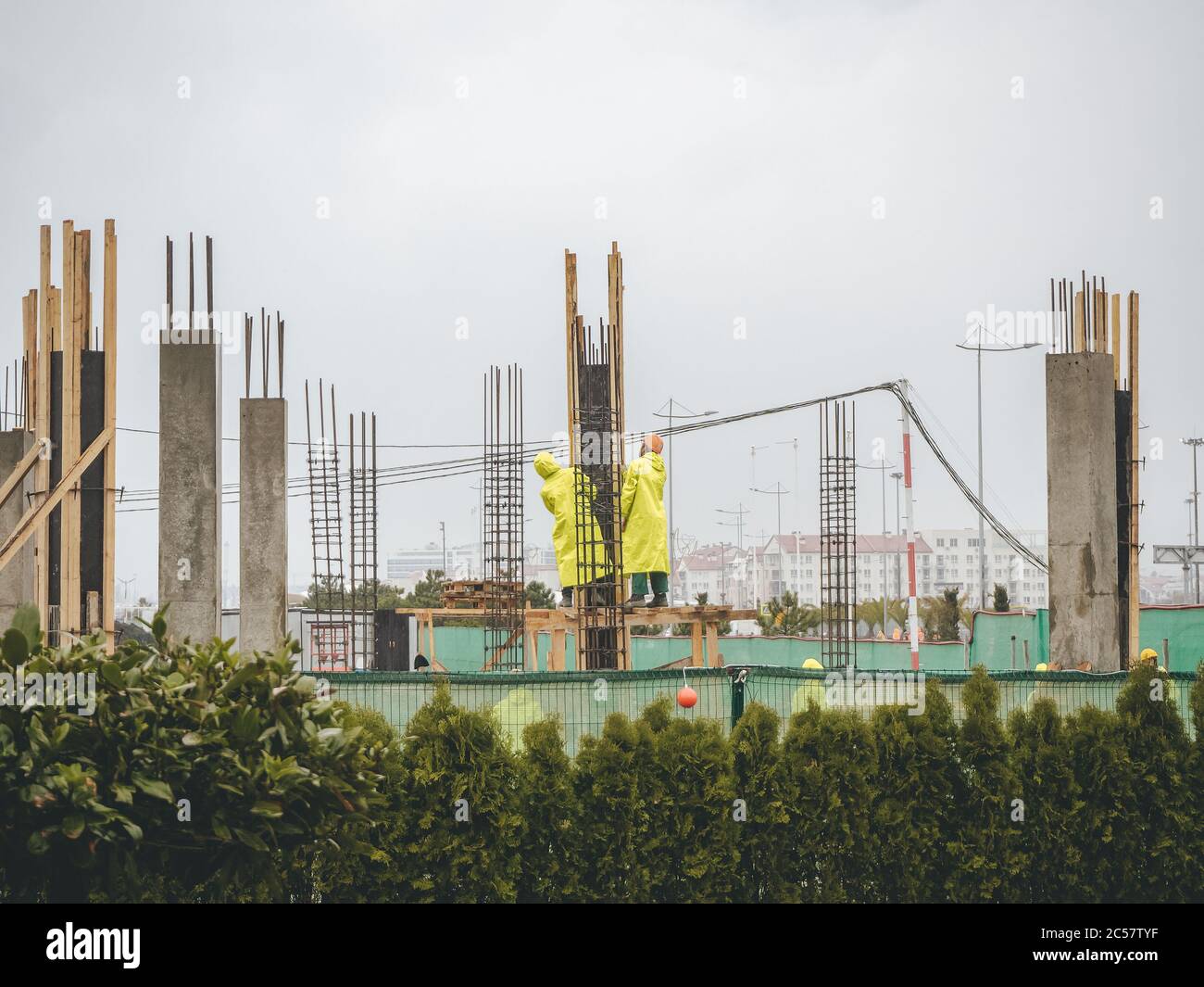 Arbeiter in gelben Regenmänteln stehen und schauen zwischen den von grünen Pflanzen umgebenen Gebäudetränen gegen einen bewölkten Himmel auf. Stockfoto