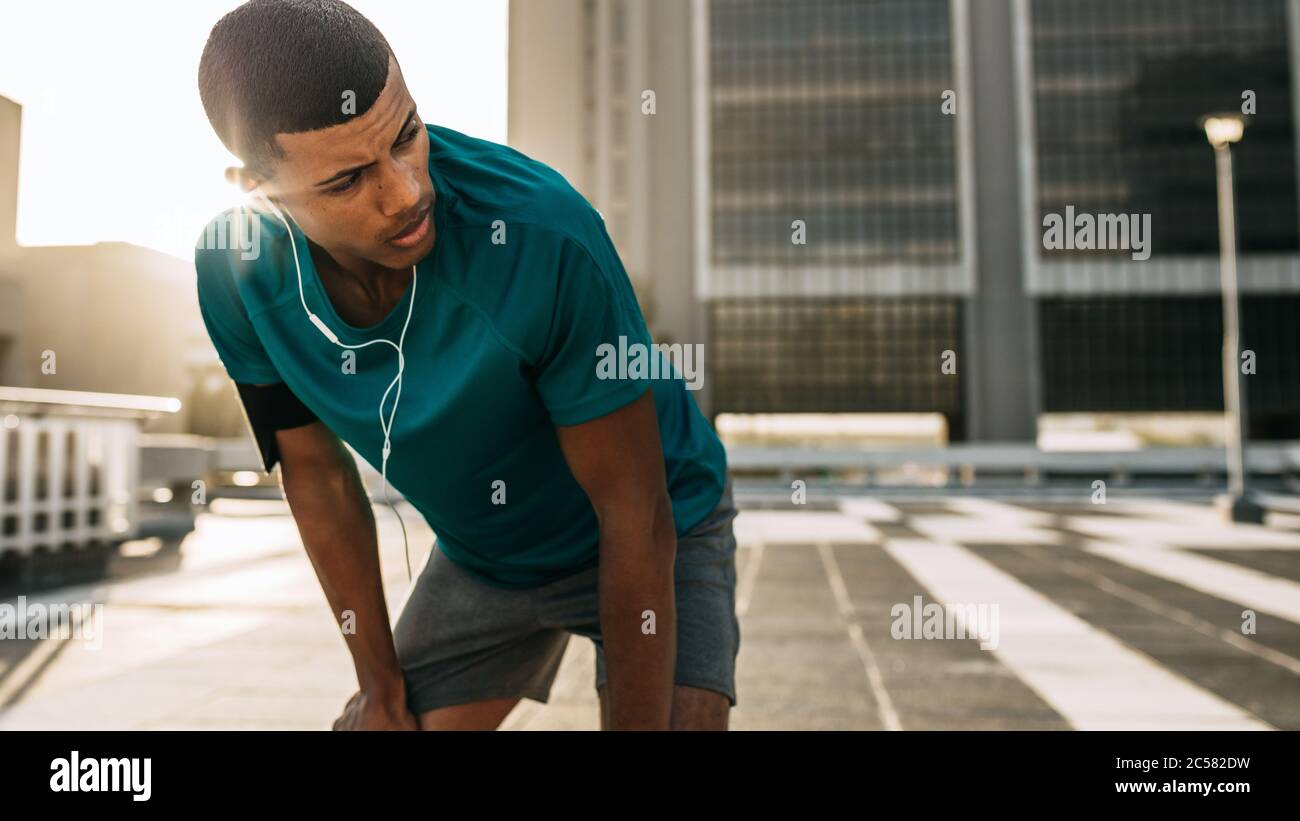 Männlicher Athlet, der sich mit den Händen auf den Knien nach vorne lehnt und in die Stadt wegschaut. Mann Läufer, die eine Pause nach dem morgendlichen Lauf. Stockfoto