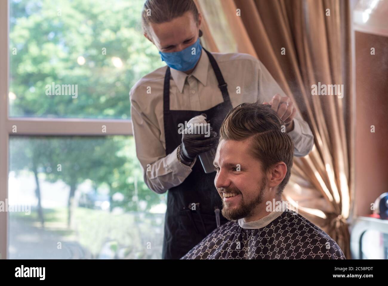 Ein Barbier in einer Schutzmaske in einem Friseursalon schneidet einen jungen bärtigen Kerl. Quarantäne Haarschnitt, Friseurmeister in einer Schutzmaske rasieren einen Kerl Stockfoto