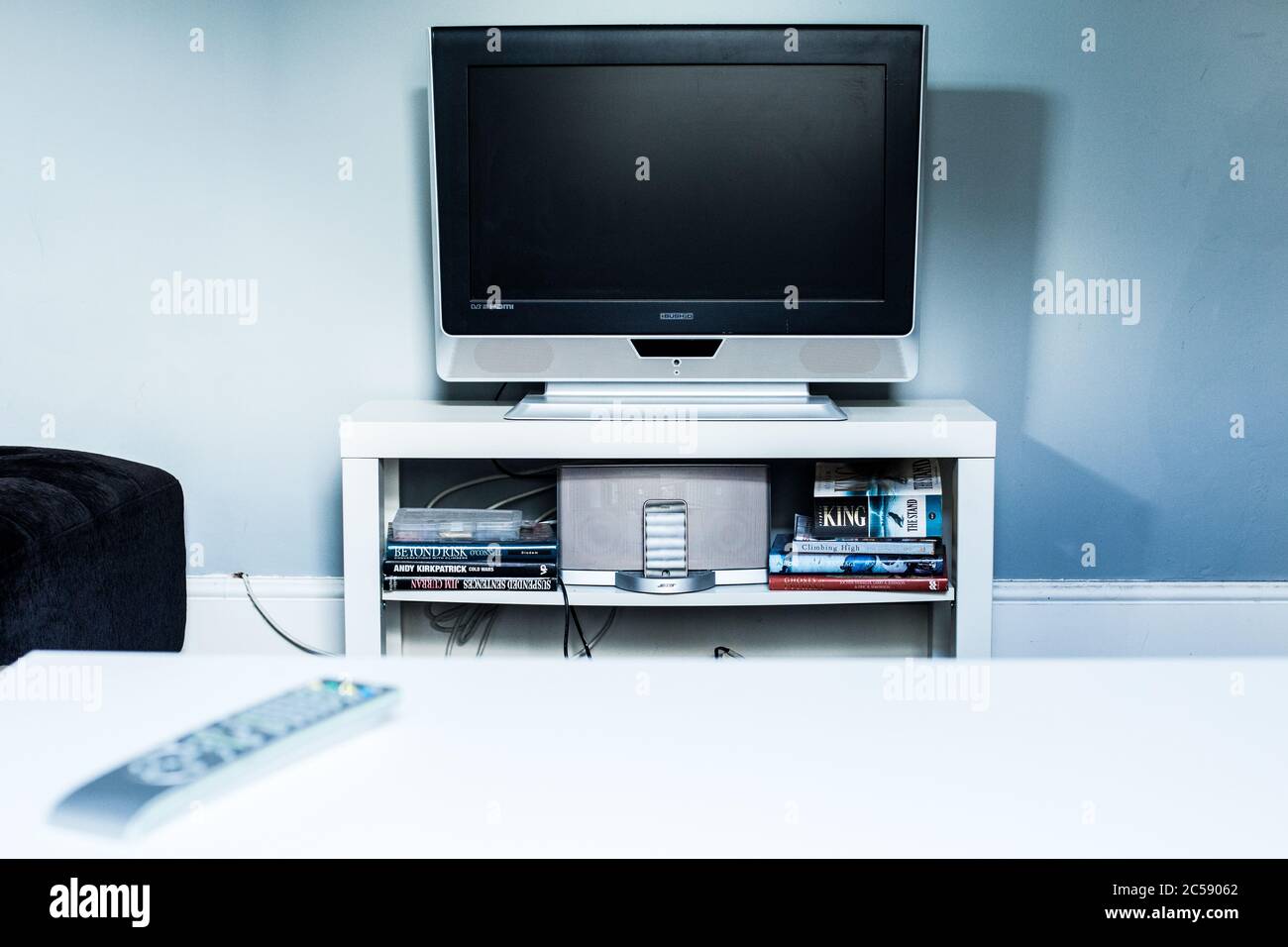 Bild eines älteren Fernsehers auf einem weißen Ständer in einem einfachen Zimmer. Es gibt einen weißen Tisch, eine Fernbedienung, Bücher und ein altes Telefon im Bild. Stockfoto
