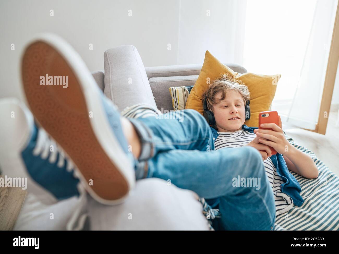Der junge Teenager liegt zu Hause auf einem gemütlichen Sofa, in lässigen Jeans und neuen Sneakers, hört Musik und chattet mit kabellosen Kopfhörern, die mit verbunden sind Stockfoto