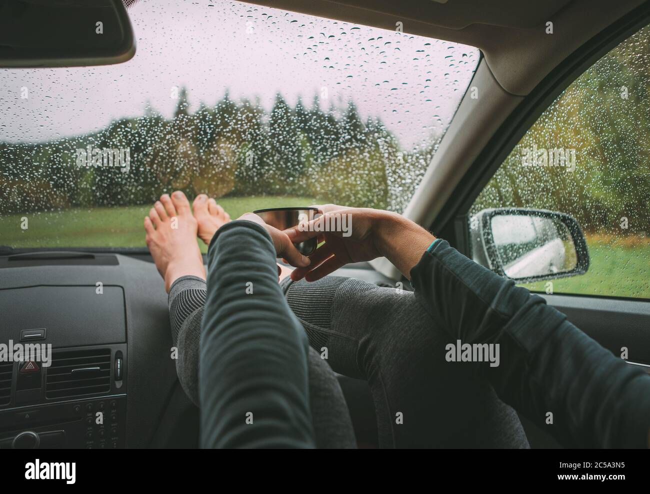 Anmutige weibliche Handflächen halten die heißen Tee Thermoskanne. Sie sitzt auf Beifahrersitz im modernen Auto, legte Beine auf Armaturenbrett genießen die launische r Stockfoto