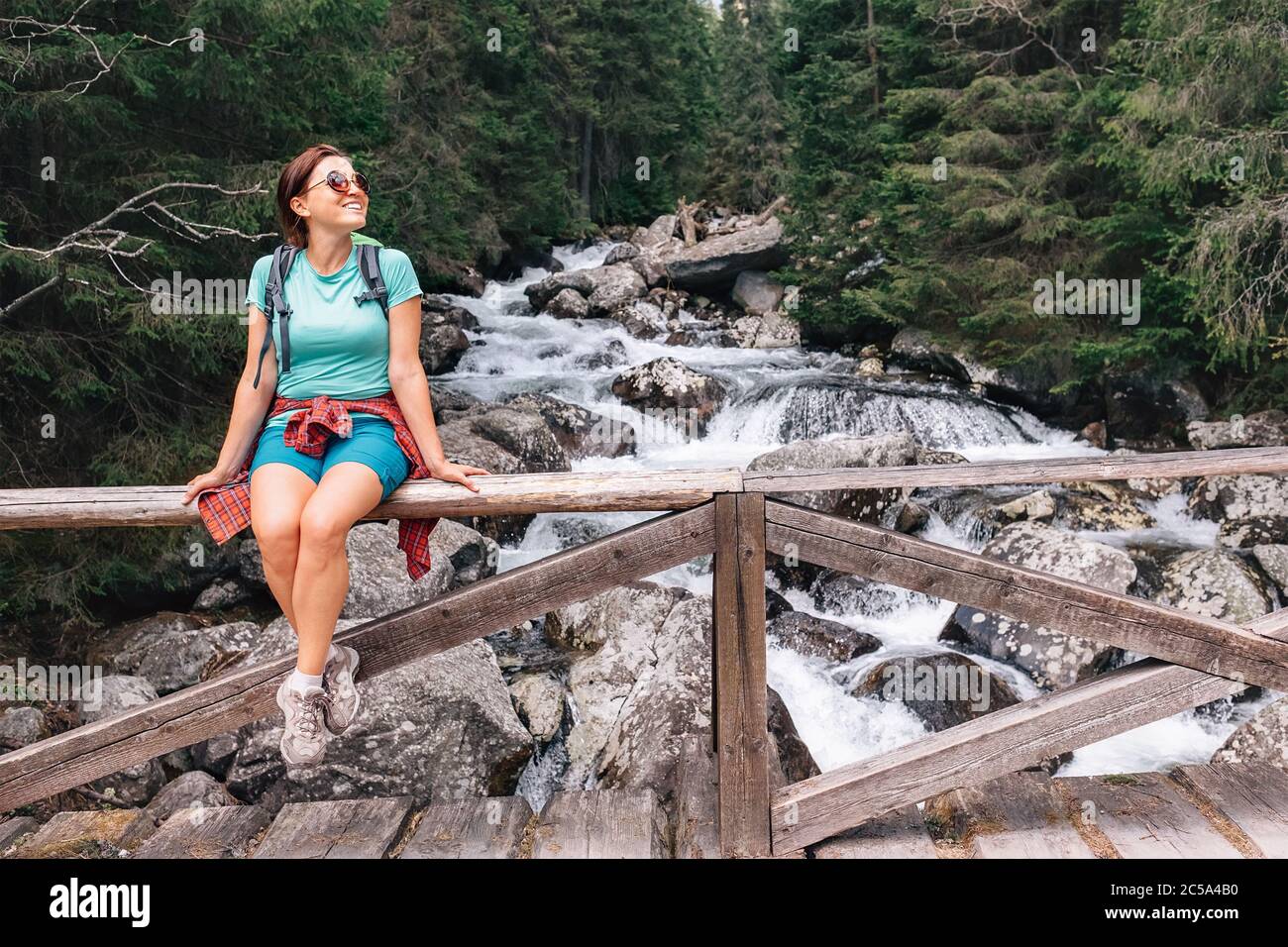 Junge Backpacker-Frau auf der Berg Kaltwasser-Bach-Brücke ruhen. Frau sitzt auf Brückenzaun lächelnd und genießen den entspannenden Moment. Acti Stockfoto