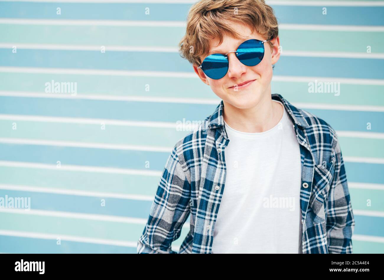 Blonde Haare 12 Jahre alten kaukasischen Teenager junge Mode-Portrait gekleidet weißen T-Shirt mit karierten Hemd in blauer Sonnenbrille mit türkisblauen Rücken Stockfoto